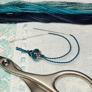 May include: A close-up of embroidery supplies, including teal and navy blue thread, a needle with teal thread, a decorative bead, and silver scissors. The supplies are arranged on a light blue and white patterned fabric.