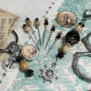 May include: A silver charm with an anchor, a ship, and a wheel, with brown and black beads, and silver pins with black and brown tops. The charm is attached to a silver key ring.