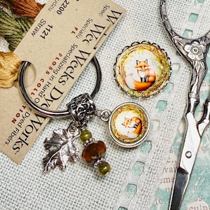 May include: A collection of craft supplies including embroidery floss, a keychain with charms, and decorative scissors. The keychain features a leaf charm, beads, and two round pendants with a fox illustration. The floss is labeled "Straw" and "Udzu".