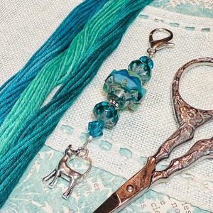 Puede incluir: Primer plano de suministros de artesanía que incluyen hilo de bordar verde azulado, unas tijeras decorativas y un dije con cuentas con un colgante de ciervo plateado. El dije presenta cuentas de vidrio azules y transparentes. Las tijeras tienen un diseño floral.
