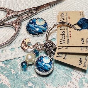 May include: A collection of craft supplies including embroidery floss, scissors, and decorative charms. The charms feature a moon and star design with blue and white accents. The floss is a deep navy blue.