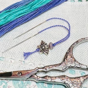 May include: A close-up of embroidery supplies including two needles, a pair of ornate scissors, and a length of purple and teal embroidery floss with a silver flower charm attached.
