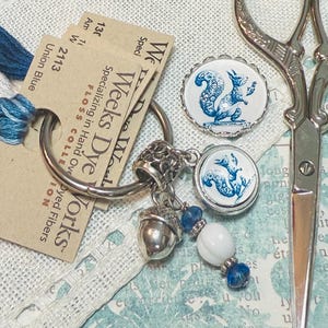 May include: A silver keyring with a blue and white squirrel charm, a silver acorn charm, and blue and white beads. The keyring is on a white fabric background with a label that reads "Weeks Dye Works" and "Specializing in Hand Dyed Fibers".