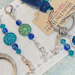 Puede incluir: Primer plano de accesorios de artesanía que incluyen dos marcadores de puntada con cuentas con dijes de sirena, un llavero con un dije de sirena y un par de tijeras. Los marcadores de puntada presentan cuentas azules y turquesas. El texto en la etiqueta dice "Weeks Dye Works".