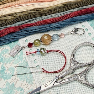 May include: A collection of embroidery supplies, including colorful thread skeins, a beaded charm with a lobster clasp, a needle threader, and ornate embroidery scissors. The threads are in shades of green, red, and beige.