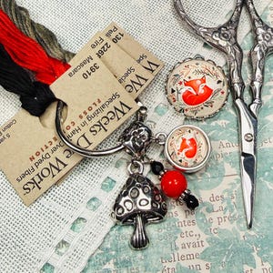 May include: An assortment of craft supplies including embroidery floss in black, red, and olive green, Weeks Dye Works floss cards, a mushroom charm, fox-themed buttons, and ornate scissors. The image showcases various crafting elements.