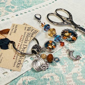 May include: A collection of craft items: embroidery floss, a charm bracelet with pumpkin and bead details, a decorative pin with a pumpkin design, and vintage scissors. The floss is labelled "We Weeks Dye Works".