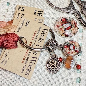 May include: A close-up of a silver scissor charm with two snowman charms, a snowflake charm, and a brown bead. The charm is attached to two spools of embroidery floss, one red and one beige, with the label "Weeks Dye Works" and the floss numbers "1330" and "225".