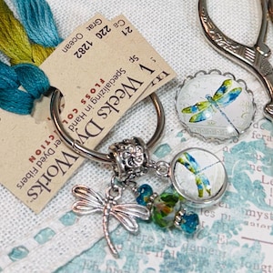 Puede incluir: Una colección de materiales de manualidades que incluye hilo de bordar turquesa y verde amarillento, un dije de libélula y tijeras decorativas. El dije presenta una libélula plateada y dos colgantes redondos con diseños de libélulas. Las tijeras son plateadas.