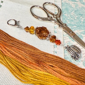 May include: A close-up of a colorful embroidery floss organizer with a pumpkin-shaped charm and a pair of silver scissors. The floss is arranged in shades of orange, yellow, and brown.