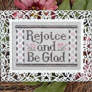May include: White framed cross-stitch embroidery with the text "Rejoice and Be Glad" in brown thread. The embroidery is surrounded by a white decorative frame with floral designs.
