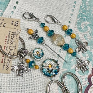 May include: A set of bee-themed needle minders and a pair of small scissors. The needle minders feature a clear glass bead, blue and yellow glass beads, and a silver bee charm. The scissors have a silver handle and a silver blade. The background is a white fabric with a floral pattern.