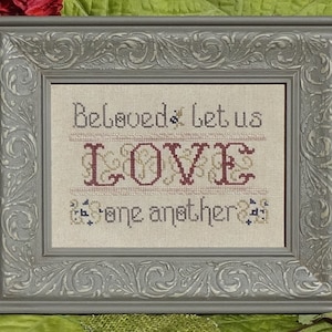 May include: A framed cross-stitch embroidery with the text "Beloved, let us LOVE one another". The embroidery is done in red, gold, and blue thread on a beige linen fabric. The frame is a gray ornate wood frame.