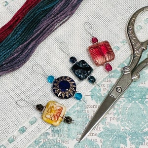 May include: Four colorful glass bead needle threaders with silver metal loops. The beads are in shades of red, blue, and gold. The threaders are on a white fabric background with a pair of silver scissors.
