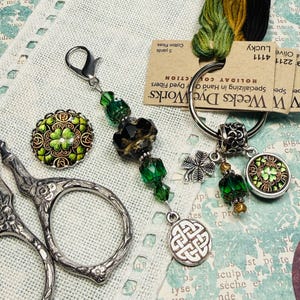 May include: A collection of craft supplies including a beaded scissor fob with green beads and a Celtic knot charm, a decorative button with a shamrock design, and embroidery floss. Silver scissors are also visible.