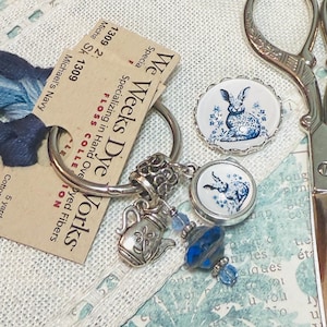 May include: A silver key chain with a teapot charm, blue glass beads, and two round charms with a blue and white rabbit design. The charms are attached to a silver ring. The key chain is on a white fabric background with a tag that reads "Weeks Dye Works" and "Specializing in Hand Dyed Fibers".