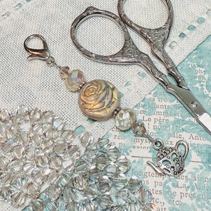 Puede incluir: Primer plano de un dije decorativo plateado con una cuenta en forma de rosa, cuentas de cristal transparente y un dije de tetera. Tijeras plateadas en el fondo.