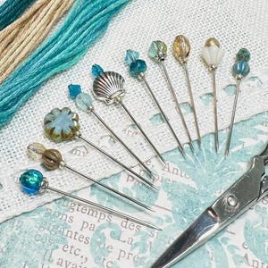 May include: A collection of decorative sewing pins with various bead designs, including a seashell, flower, and faceted beads. Teal and tan embroidery floss and vintage scissors are also in the image. The items are arranged on a patterned paper background.