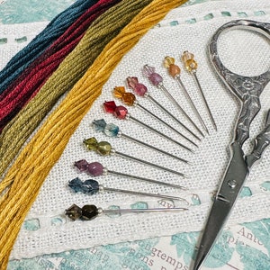 May include: A set of 10 colourful glass-headed pins with silver needles, arranged in a fan shape on a white fabric background.  There are also four spools of embroidery floss in various shades of brown, red, and blue.