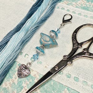 Puede incluir: Imagen de temática artesanal con hilo de bordar azul, un charm decorativo de pájaro, un charm de corazón y unas tijeras ornamentadas. El hilo es en tonos azules. El charm de pájaro es turquesa y plateado. Las tijeras son plateadas.