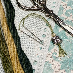 Puede incluir: Un surtido de materiales de bordado, que incluye hilos verdes y dorados, tijeras plateadas y un sujetador de agujas con un dije de conejito. Los artículos están dispuestos sobre un fondo estampado, sugiriendo un tema de manualidades.