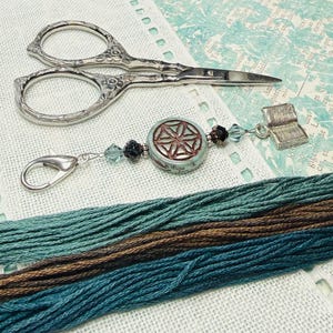 May include: A collection of craft supplies including silver embroidery scissors, a decorative charm with a starburst design, and a small book charm. Several skeins of teal and brown embroidery floss are also present, suggesting a needlework theme.