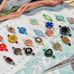 May include: A set of 18 colorful glass needle threaders with different designs, including flowers, hearts, and geometric shapes. The threaders are arranged on a white fabric surface with a pair of silver scissors.
