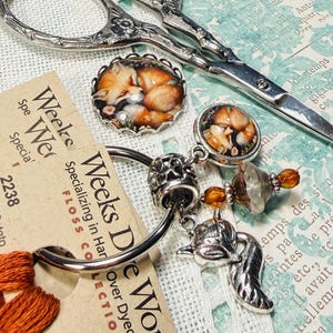 Könnte beinhalten: Eine Sammlung von Bastelmaterialien, darunter Stickgarn, ein Metallarmband mit Fuchsanhängern, eine silberne Schere und Garnkarten. Das Garn ist in Orange- und Beigetönen gehalten.