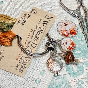 May include: A silver key chain with a variety of charms, including a small sheep, a brown bead, and three round charms with images of pumpkins and autumn foliage. The key chain is attached to a card with the text "W' Weeks Dye Works" and "Specializing in Hand Dyed Fibres".