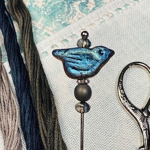 May include: A handcrafted blue bird pin with a beaded stem, set against a backdrop of embroidery floss in shades of blue, gray, and brown. A pair of silver scissors is also visible.