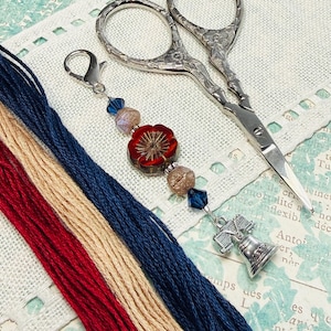 May include: A craft-themed image featuring red, beige, and navy blue embroidery floss, a silver charm with a Liberty Bell design, and ornate scissors. A beaded charm with a red flower and blue crystal accents is also present.