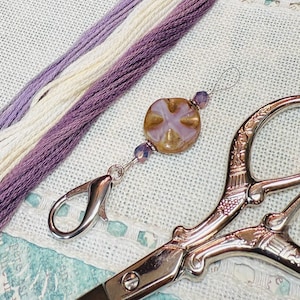 Puede incluir: Primer plano de suministros de manualidades que incluyen hilo de bordar en tonos morados y crema, un adorno decorativo para tijeras con una cuenta en forma de cruz y tijeras plateadas ornamentadas. Los artículos están dispuestos sobre una superficie clara y texturizada.