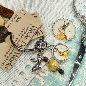 以下が含まれることがあります： Weeks Dye Worksの刺繍糸、ウサギのチャームが付いたシルバーのチャームブレスレット、ウサギをテーマにした2つのカボションなど、さまざまなクラフト用品。ハサミと刺繍糸も写っています。