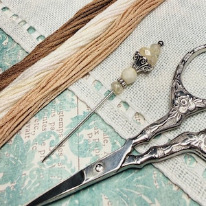 May include: An assortment of embroidery supplies, including three skeins of thread in shades of brown and cream, a decorative needle with a beaded top, and ornate silver scissors. The items are arranged on a patterned fabric.