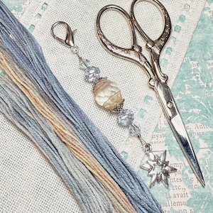 Puede incluir: Un marcador de punto plateado y blanco con un encanto de estrella, un ovillo de hilo de bordar azul y beige y un par de tijeras de bordar plateadas sobre un fondo de lino blanco.