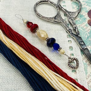 May include: A close-up of embroidery supplies including red, cream, and navy blue thread, a beaded needle minder, and ornate silver scissors. The needle minder has a heart-shaped charm and colorful beads.