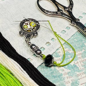 May include: A needle minder with a floral design, a black bead, and green thread, alongside embroidery floss and scissors. The needle minder has a silver-colored metal base with a decorative floral pattern in green, yellow, and white.