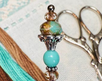 May include: A decorative needle minder with a silver-colored base and a teal and brown beaded design. The needle minder has a round teal bead, a faceted teal bead, and a small silver-colored bead at the top. A pair of silver scissors and embroidery floss are in the background.