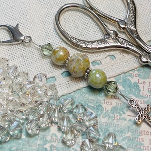 Puede incluir: Un charm de tijera plateado con una cuenta de piedra verde y marrón, una cuenta de cristal transparente y un charm de cruz plateado. El charm está unido a un anillo plateado.