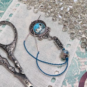 May include: A silver-toned needle minder with a blue and white bird design, a needle, and blue thread rest on white fabric. Decorative floral scissors and scattered crystals complete the craft-themed image.