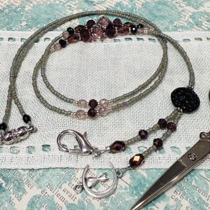 May include: A beaded lanyard with a silver clasp and a black decorative bead. The lanyard is gray with purple and clear beads. A pair of silver scissors is also in the image.