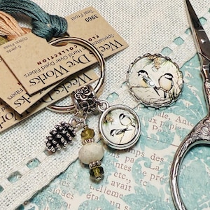 May include: A collection of craft supplies including Weeks Dye Works cotton floss, a charm bracelet with bird-themed pendants, a decorative button, and ornate scissors. The items are arranged on a patterned background with lace trim.