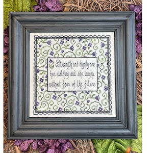 Puede incluir: Un bordado de punto de cruz enmarcado con fondo blanco y un patrón floral verde y morado. El texto dice "Strength and dignity are her clothing and she laughs without fear of the future."