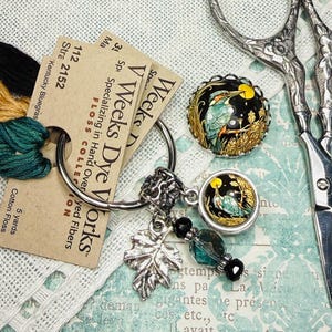 May include: An assortment of craft supplies including embroidery floss in black, gold, and green, a pair of silver scissors, and decorative buttons with a bird design. Also included are tags from V. Weeks Dye Works.