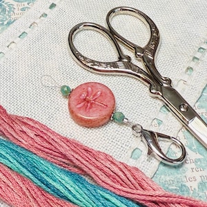 Puede incluir: Primer plano de suministros de bordado que incluyen tijeras plateadas, hilo de bordar rosa y turquesa, y un dije de libélula rosa con cuentas verdes. Los artículos están dispuestos sobre una tela blanca.