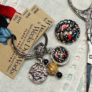 May include: A collection of craft supplies including embroidery floss in red, black, and blue, a decorative button with a fox design, and a pair of ornate scissors. The image also shows a charm with a floral design and a metal ring.