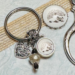May include: Silver key ring with a heart charm, a crystal bead, and two round glass charms featuring a hedgehog illustration.