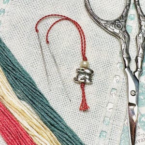 May include: A close-up of a red embroidery floss with a silver rabbit charm and a pearl bead. Two needles are also visible on a white linen fabric.