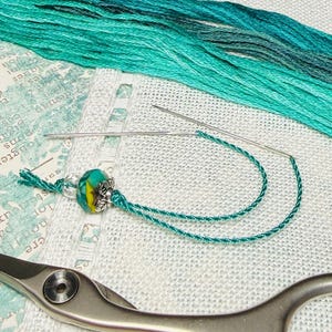 Puede incluir: Primer plano de suministros de bordado. Una aguja enhebrada con hilo verde azulado está junto a una borla con cuentas. También se ven hilo de bordar verde azulado y un par de tijeras. El fondo es una tela de color claro.