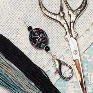 Puede incluir: Primer plano de suministros de bordado. Una cuenta decorativa con patrones arremolinados en tonos azules, negros y marrones está unida a un cierre plateado. También se ven tijeras plateadas e hilo de bordar negro, gris y verde azulado.